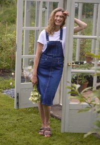 Denim midi overall klänning med justerbara remmar, framfickor och en åtsittande byst. Modellen håller en liten bukett; tillsammans med bruna sandaler.
