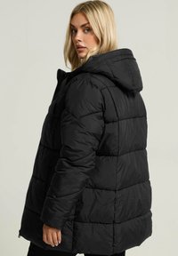 Doudoune noire avec capuche, présentant un design matelassé et une fermeture éclair à l'avant. Le matériau semble lisse et légèrement brillant.