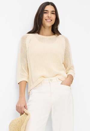 Frau lächelnd in einem cremefarbenen locker gestrickten Pullover und weißen Hosen, hält in einer Hand einen beigefarbenen gewebten Hut, steht vor einem weißen Hintergrund.