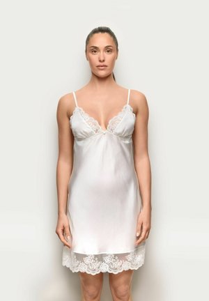 Nainen seisoo kasvot eteenpäin valkoisessa satiinisessä liivimekossa, jossa on pitsireunus pääntiellä ja helmassa, yksinkertaisella taustalla.