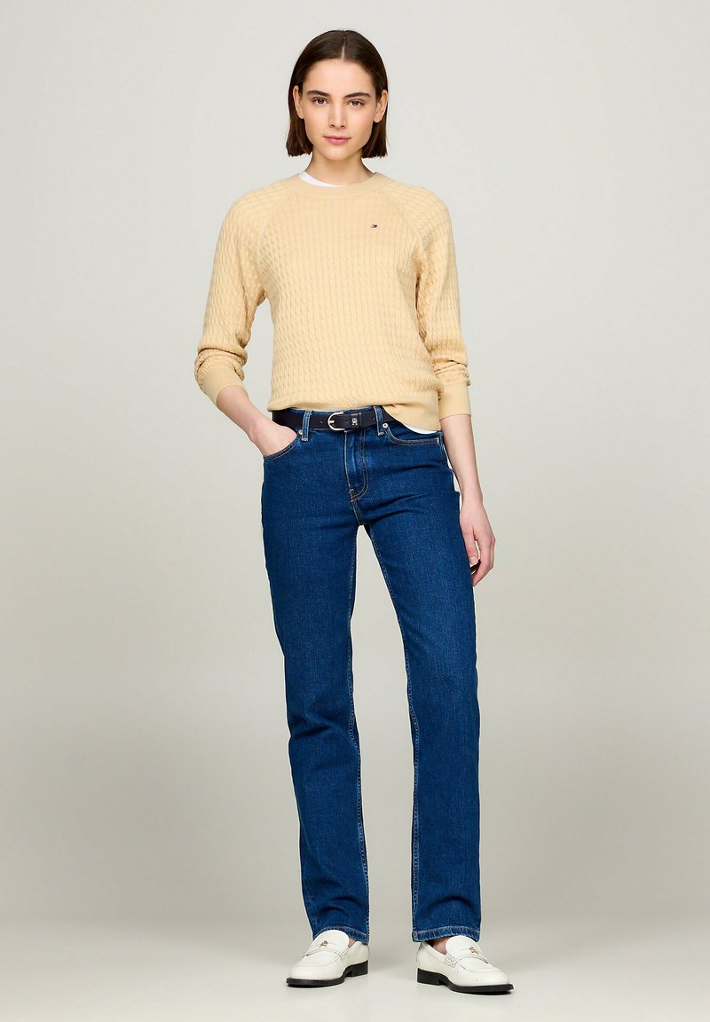 Beżowy sweter robiony na drutach z teksturowanym wzorem, w połączeniu z ciemnoniebieskimi dżinsami o prostych nogawkach i białymi butami. Modelka stoi pewnie.