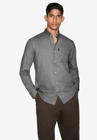 Grå button-up skjorte med en subtil tekstur, der har en lomme og afrundet kant, parret med mørkebrune bukser.