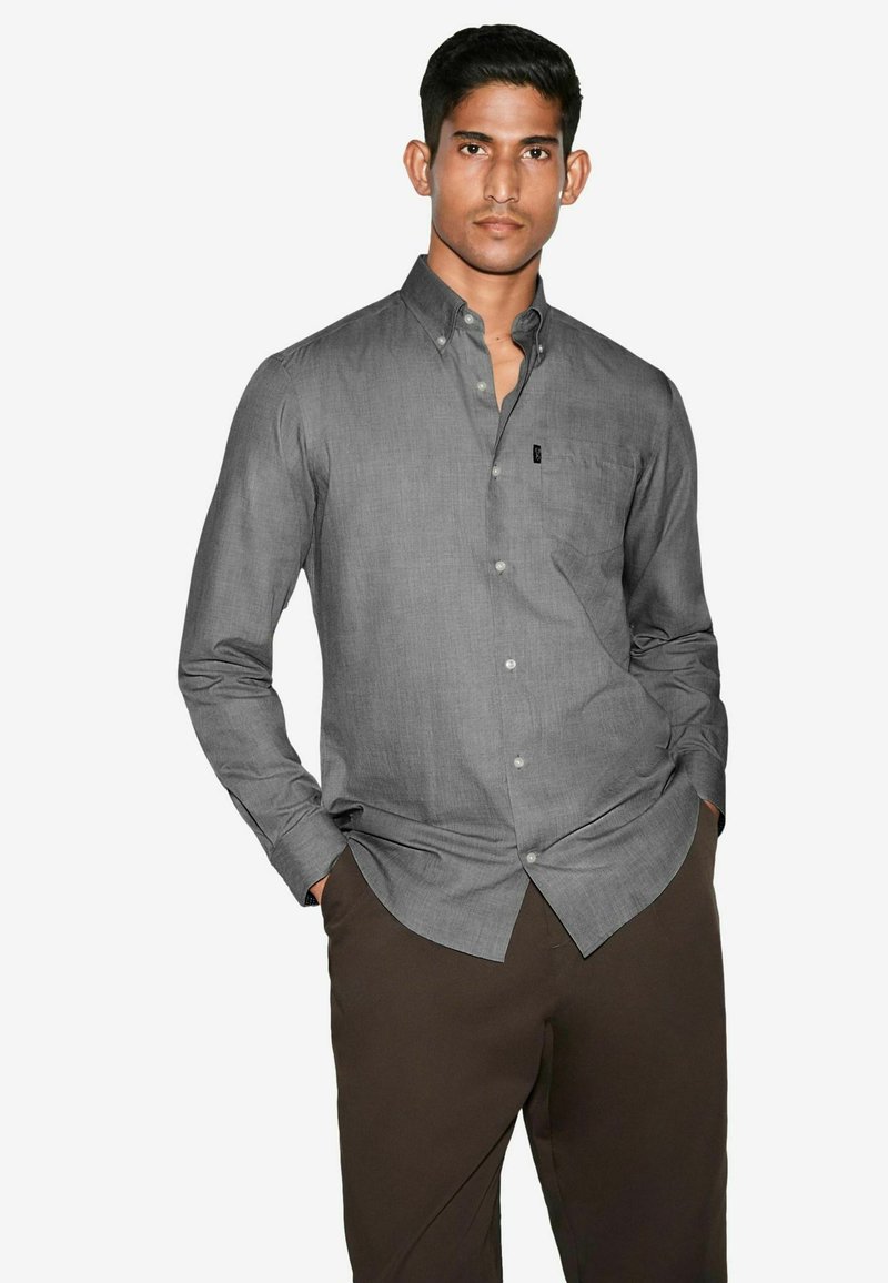 Grå button-up skjorte med en subtil tekstur, der har en lomme og afrundet kant, parret med mørkebrune bukser.