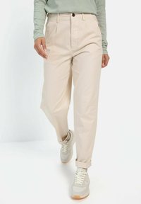 Beige bukser med løs pasform, der har frontfolder og sidelommer. Stylet med lyse sneakers og en langærmet grøn top.