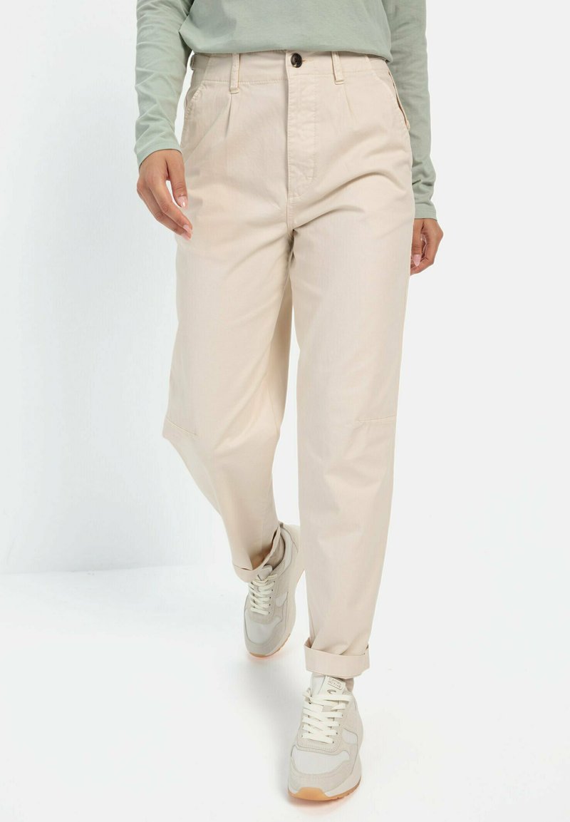 Beige bukser med løs pasform, der har frontfolder og sidelommer. Stylet med lyse sneakers og en langærmet grøn top.