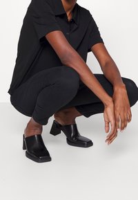 Personne accroupie, portant un haut à manches courtes noir, un pantalon noir et des mules à talons carrés noires sur un fond blanc uni.