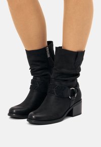 Bottines noires avec un design froissé, dotées d'une fermeture éclair sur le côté, d'une sangle texturée avec des motifs géométriques et d'un petit talon carré.