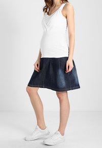 Top blanco sin mangas con diseño entrelazado, combinado con una falda de mezclilla oscura que se ensancha en el dobladillo. Los zapatos son zapatillas deportivas blancas.