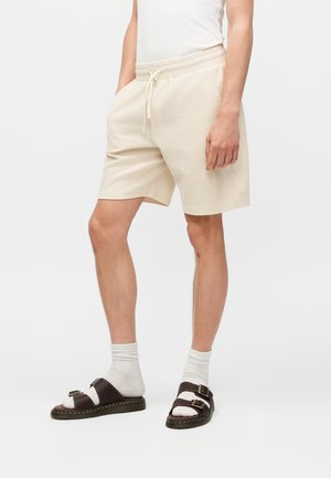 Person som har på seg beige kortbukser med tekstur, elastisk midje og snøring, hvite sokker og mørkebrune sandaler med spenne.