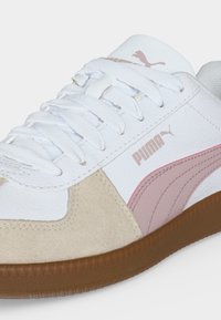 Άσπρο και μπεζ αθλητικό παπούτσι Puma με ροζ λογότυπο, άσπρα κορδόνια και καφέ λαστιχένια σόλα, δείχνοντάς πλευρικές και επάνω λεπτομέρειες.