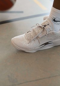 Fehér sportcipő, amely texturált felsőrésszel rendelkezik finom mintázattal, fűzőkkel és ezüst díszítéssel az oldalán. Fehér zoknival viselve.