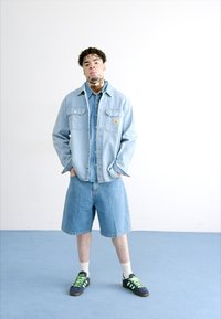 Lyseblå denimjakke, matchende shorts og sorte træningssko med grønne detaljer. Hvide sokker er synlige. Baggrunden er ensfarvet og lys.