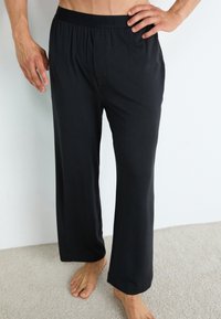 Homme portant un pantalon de lounge noir à coupe large de Calvin Klein, debout pieds nus sur un tapis contre un mur blanc.