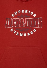 Rdeča kapuca z grafičnim dizajnom, ki vsebuje besedilo "SUPERIOR JACK & JONES" in "STANDARD" v kontrastnih barvah. Gladka tekstura, sprednji žep.