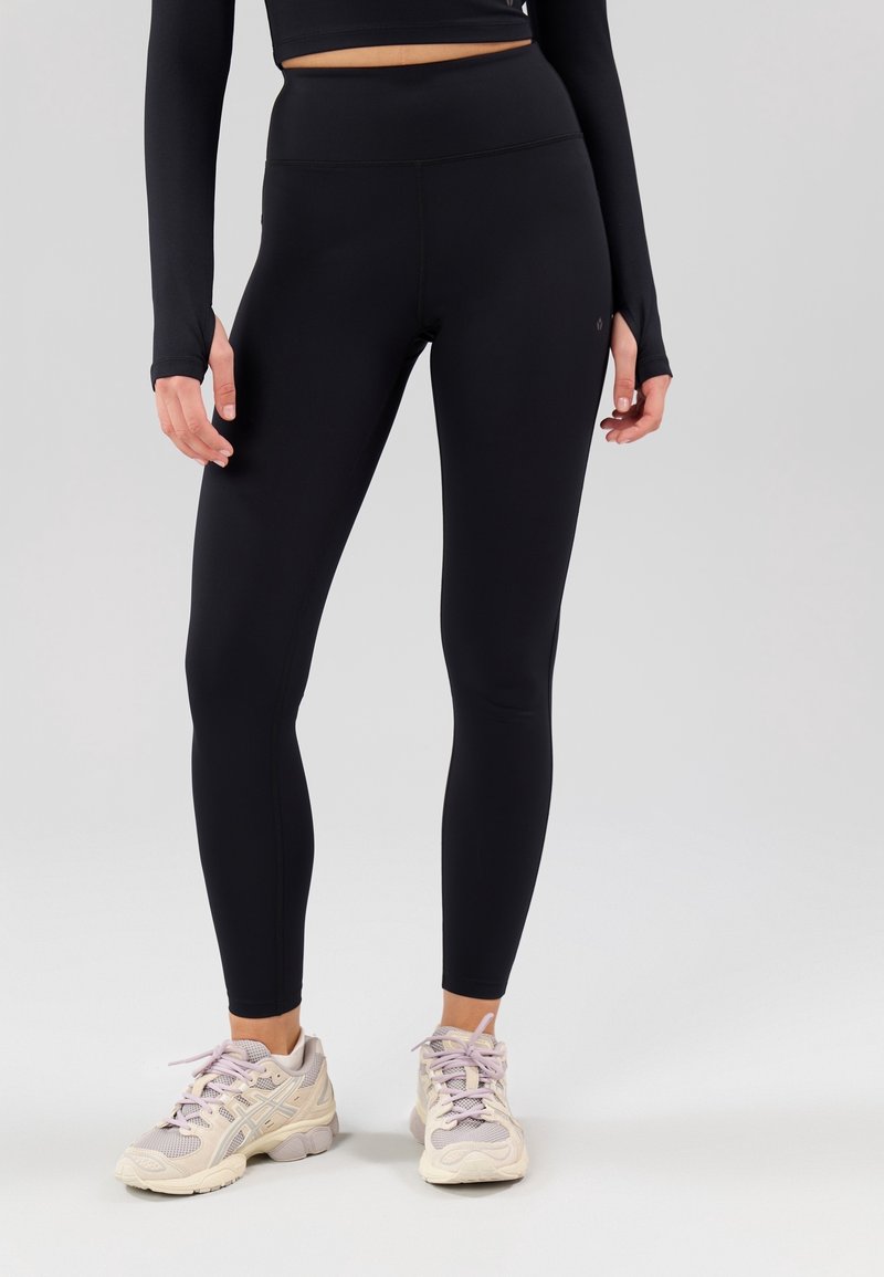 Leggings negros de talle alto con una textura suave y elástica y costuras mínimas. Combinados con zapatillas deportivas de color claro y una camiseta de manga larga.