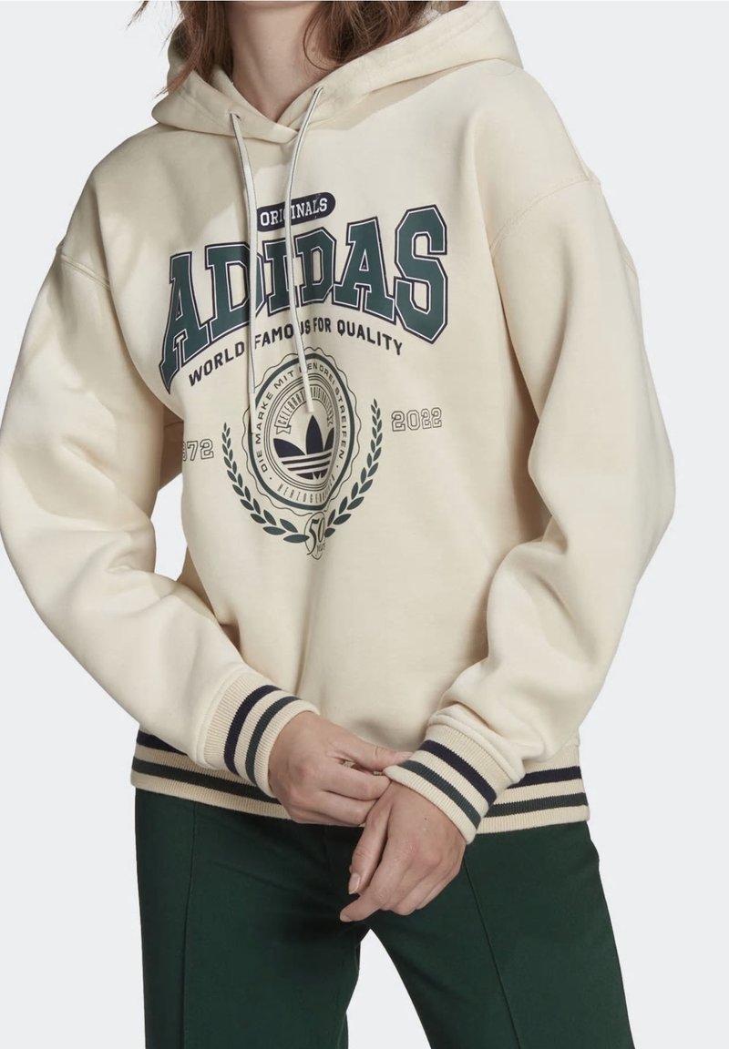 Persona che indossa una felpa Adidas crema con logo e testo verde scuro, abbinata a pantaloni verde scuro, mani incrociate in vita.