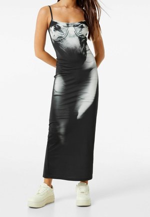 Femme portant une robe maxi débardeur dégradée noir et blanc avec des bretelles fines, associée à des baskets blanches épaisses, debout devant un fond blanc uni.