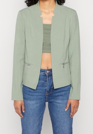 Femme portant un blazer vert clair ouvert sur un crop top assorti avec un jean bleu taille haute, vue de face, mains détendues le long du corps.