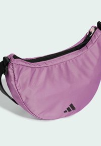 Bolso cruzado de nailon morado con forma curva, cremallera negra y logo de Adidas en negro en la esquina inferior. Textura suave, diseño ligero.