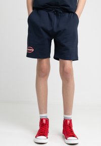 Námořnické modré bavlněné šortky s elastickým pasem a bočními kapsami. Na levém stehně je kulatý červený logo. Doplněné červenými teniskami.