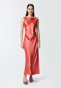 Duga, bezrukava, uska haljina u svijetloj koraljno-crvenoj boji s glatkom, sjajnom teksturom i drapiranim detaljem u struku. Uparena sa cipelama s uzorkom.