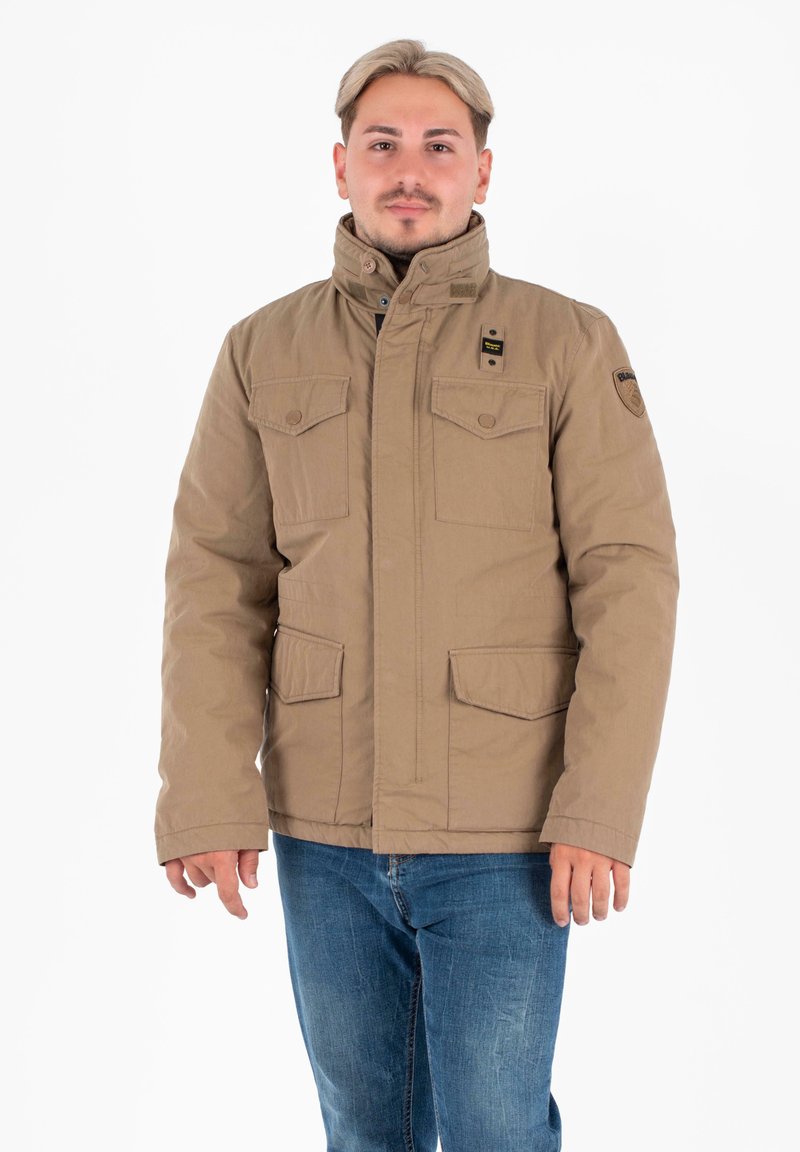 Uomo con capelli biondi corti, che indossa una giacca beige con molte tasche e jeans blu, in piedi su uno sfondo bianco.