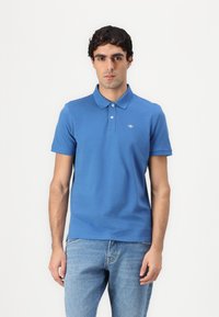 Blå bomulds polo-shirt med en to-knap lukning, korte ærmer og et lille logo på venstre bryst. Bæret sammen med lyseblå jeans.