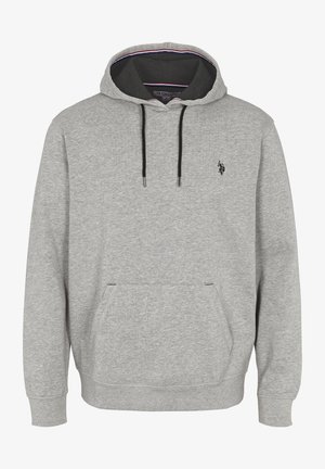 Man in een lichtgrijze hoodie met een voorzak en zwarte aantrekkoorden, gecombineerd met blauwe jeans, staande met één hand in de zak.