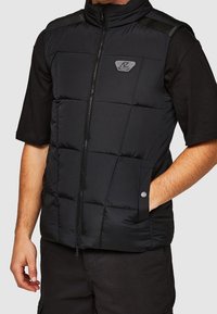 Schwarze Steppweste mit hohem Kragen, Reißverschluss vorne und Seitentaschen. Verfügt über ein gestepptes Design und ein Logoakzent auf der Schulter.