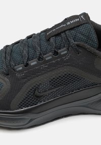 Svart og grå atletisk sko med mesh- og tekstiloverdel, rund tå, flate lisser, og en svart Nike swoosh-logo på siden.