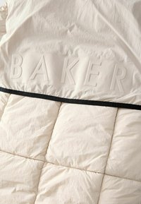 Puffasti pokrivač u krem boji s reljefnim natpisom "BAKER". Ima teksturiranu površinu i crni rub duž ivice.