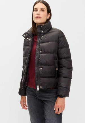 Woman wearing a black puffer jacket with snap buttons, a burgundy sweater, and dark jeans, standing against a plain light background.