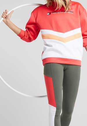 Felpa rossa e bianca con strisce a zigzag arancioni e bianche, abbinata a leggings verdi con dettagli rossi e bianchi.