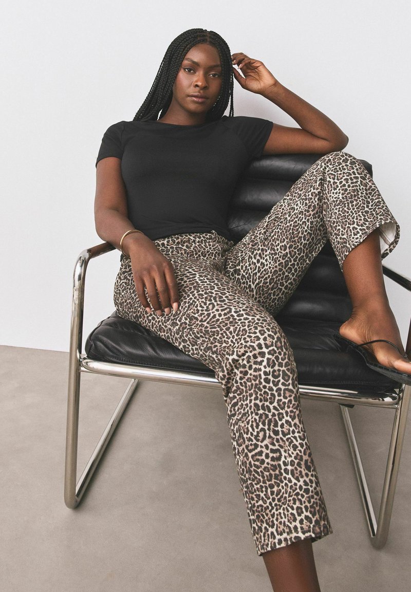 Femme avec de longues tresses portant une chemise noire et un pantalon imprimé léopard, assise détendue sur un fauteuil en cuir noir avec une structure métallique.