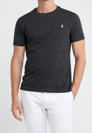 Homme portant un t-shirt gris foncé à manches courtes et col rond avec un petit logo blanc brodé sur la poitrine, ainsi qu'un pantalon blanc.