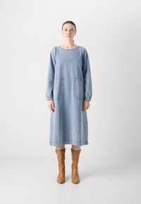 Denimkleid in Hellblau, mit langen Ärmeln, runder Ausschnitt, zwei großen Vordertaschen und gerafften Manschetten. Getragen mit beigen Stiefeletten.