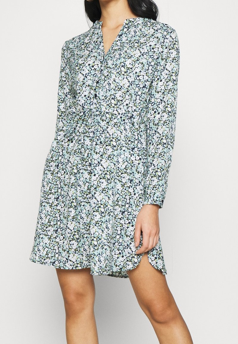 Femme portant une robe à manches longues à imprimé floral, col en V et taille resserrée, debout devant un fond clair uni.