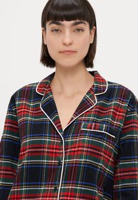Rød og grøn tartan pyjamatop med marineblå bund, hvid piping, krave og enkelt brystlomme. 100% bomuldsmateriale, knaplukning.
