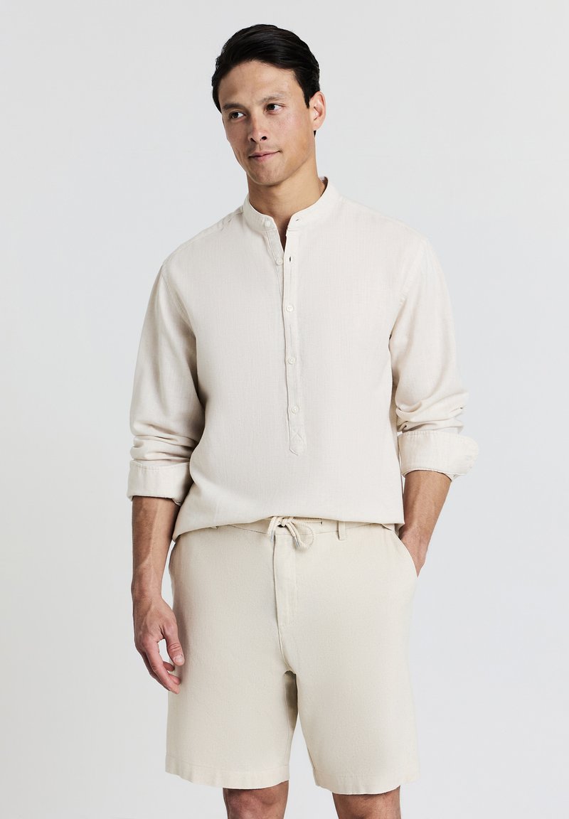 Homme portant une chemise beige à manches longues boutonnée et un short assorti, se tenant avec une main dans sa poche devant un fond uni.