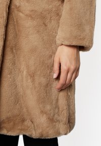 Płaszcz z futerka ekologicznego w jasnobrązowym kolorze o miękkiej, pluszowej teksturze. Charakteryzuje się luźnym krojem, mankietowanymi rękawami i gładkim wykończeniem.