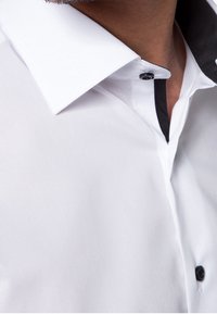 Camicia bianca con colletto interno nero e patta dei bottoni. Presenta un colletto a punta e una superficie liscia. I bottoni sono neri.