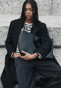 Borsa a spalla in pelle nera con texture liscia, forma angolare e design minimalista. Indossata da una persona con un cappotto nero e una maglietta a righe.