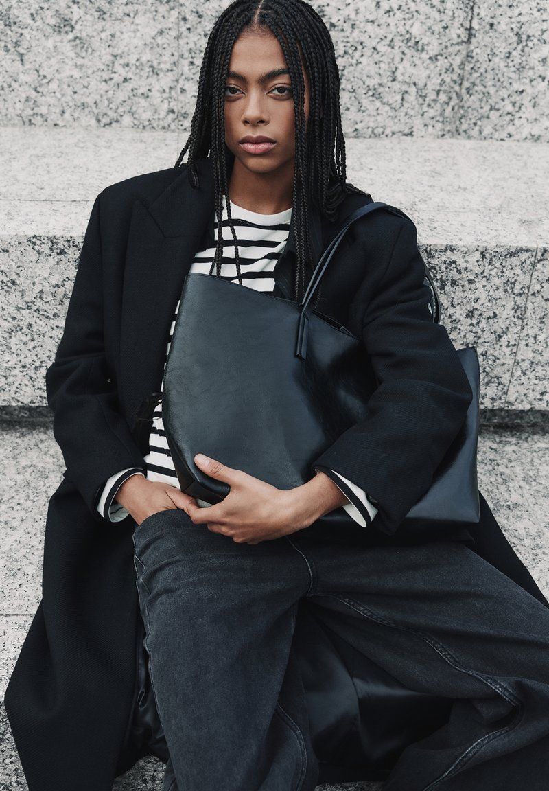 Borsa a spalla in pelle nera con texture liscia, forma angolare e design minimalista. Indossata da una persona con un cappotto nero e una maglietta a righe.