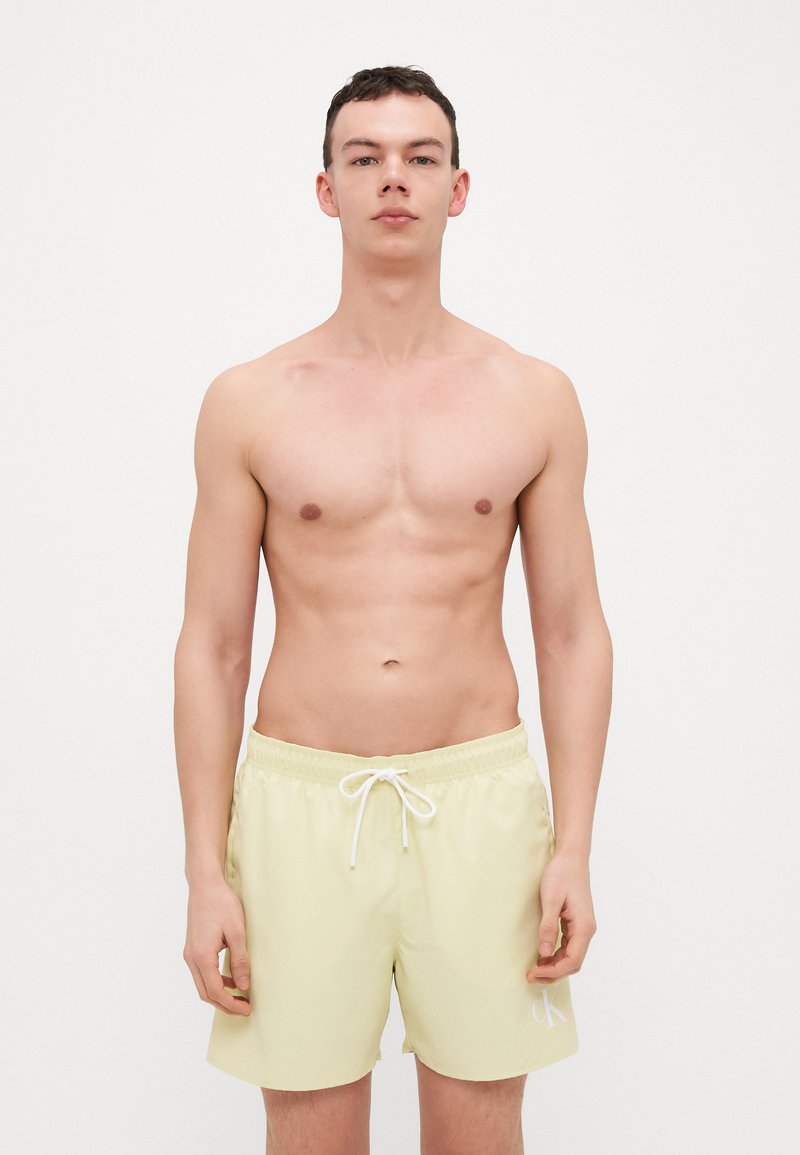 Shorts de bain pour hommes en jaune clair, fabriqués en tissu lisse, dotés d'une taille élastique avec cordon de serrage et d'un petit logo blanc sur le côté gauche.