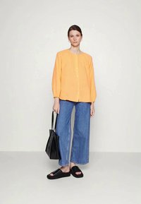 Camisa laranja de botões com decote redondo e mangas longas, combinada com jeans de ganga azul de perna larga e chinelos pretos. Uma bolsa tote preta está a ser segurada.