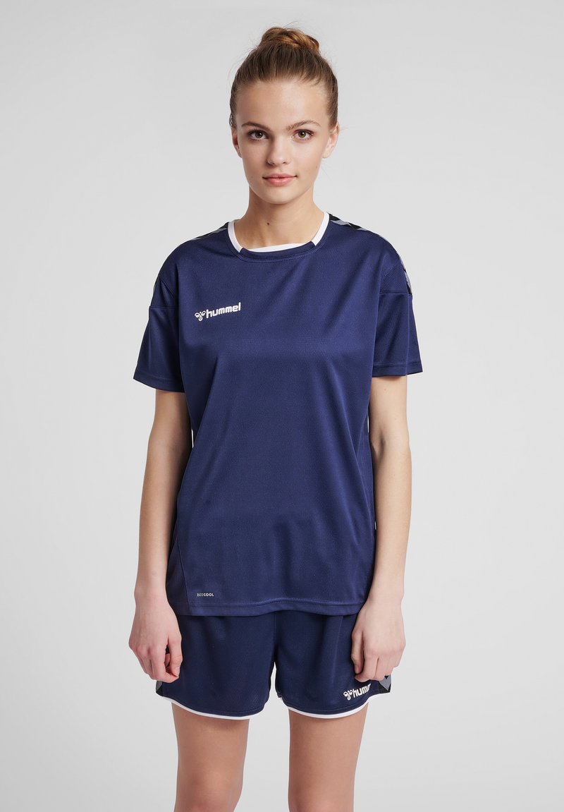Jonge vrouw met haar in een knot, gekleed in een marineblauw sportshirt en bijpassende shorts, staand tegen een effen achtergrond.