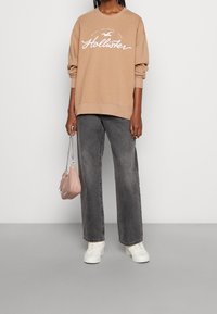 Ljusbrun sweatshirt med vit text, matchad med grå vida jeans. Slät textur, vita sneakers och en liten rosa handväska.