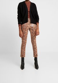 Person, die braune Schlange-Muster-Hosen mit Frontschlitzen, schwarze Knöchel-Stiefel, eine schwarze Strickjacke und ein braunes langärmliges Oberteil trägt und steht.