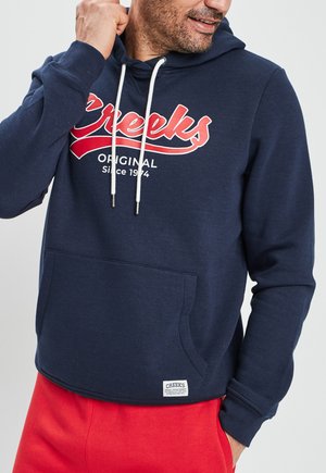 Haut à capuche bleu marine avec une poche kangourou, cordons blancs, et un texte rouge qui indique "Creeks Original Since 1974" sur le devant.