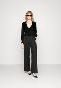 Blusa nera di satin a maniche lunghe con scollatura a V e dettaglio nodo, abbinata a pantaloni larghi in charcoal a vita alta e décolleté neri a punta.